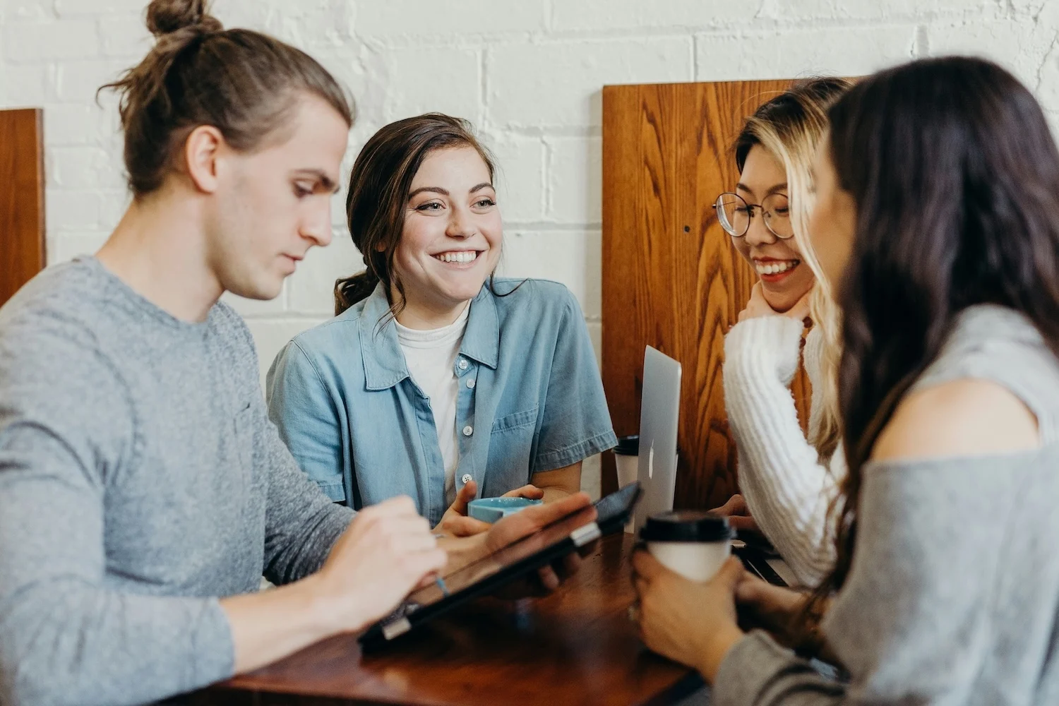 Four people sitting around a wooden table in a café, smiling and talking together, with laptops, a tablet, and coffee cups in front of them.