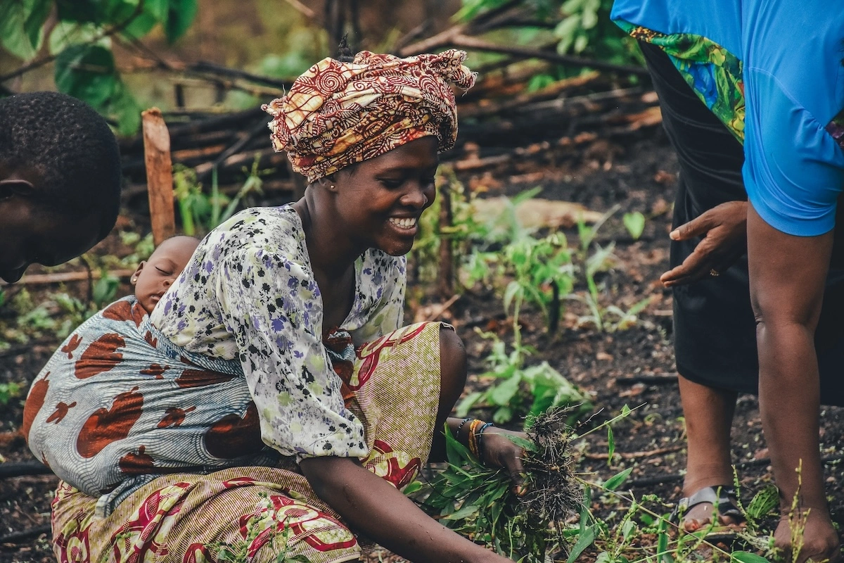 woman smiling while working the soil in a garden, carrying a sleeping baby wrapped on her back, with another person crouched nearby.