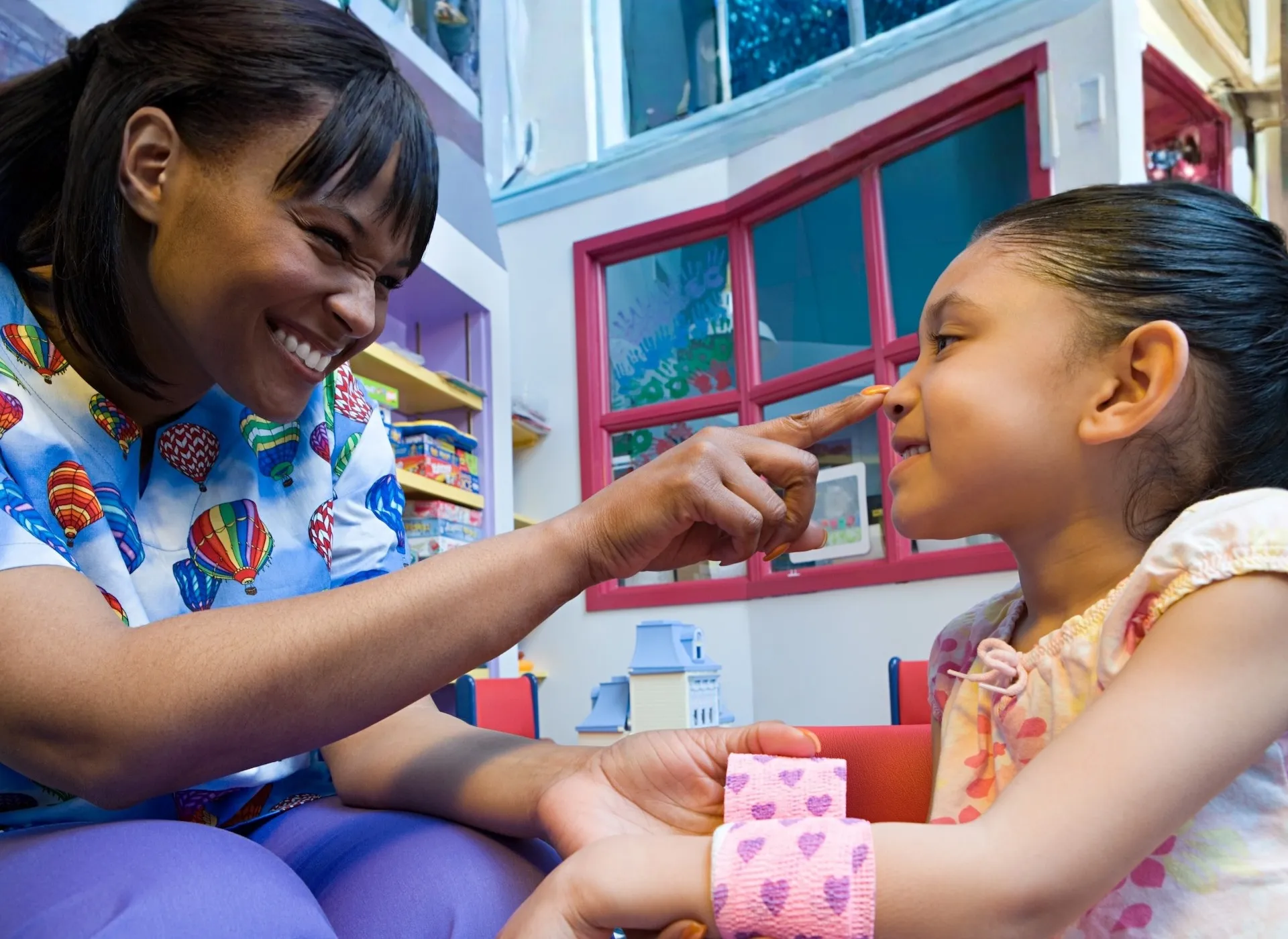 A smiling nurse gently touches a young girl’s nose while holding her arm, creating a warm, caring moment in a brightly coloured playroom.