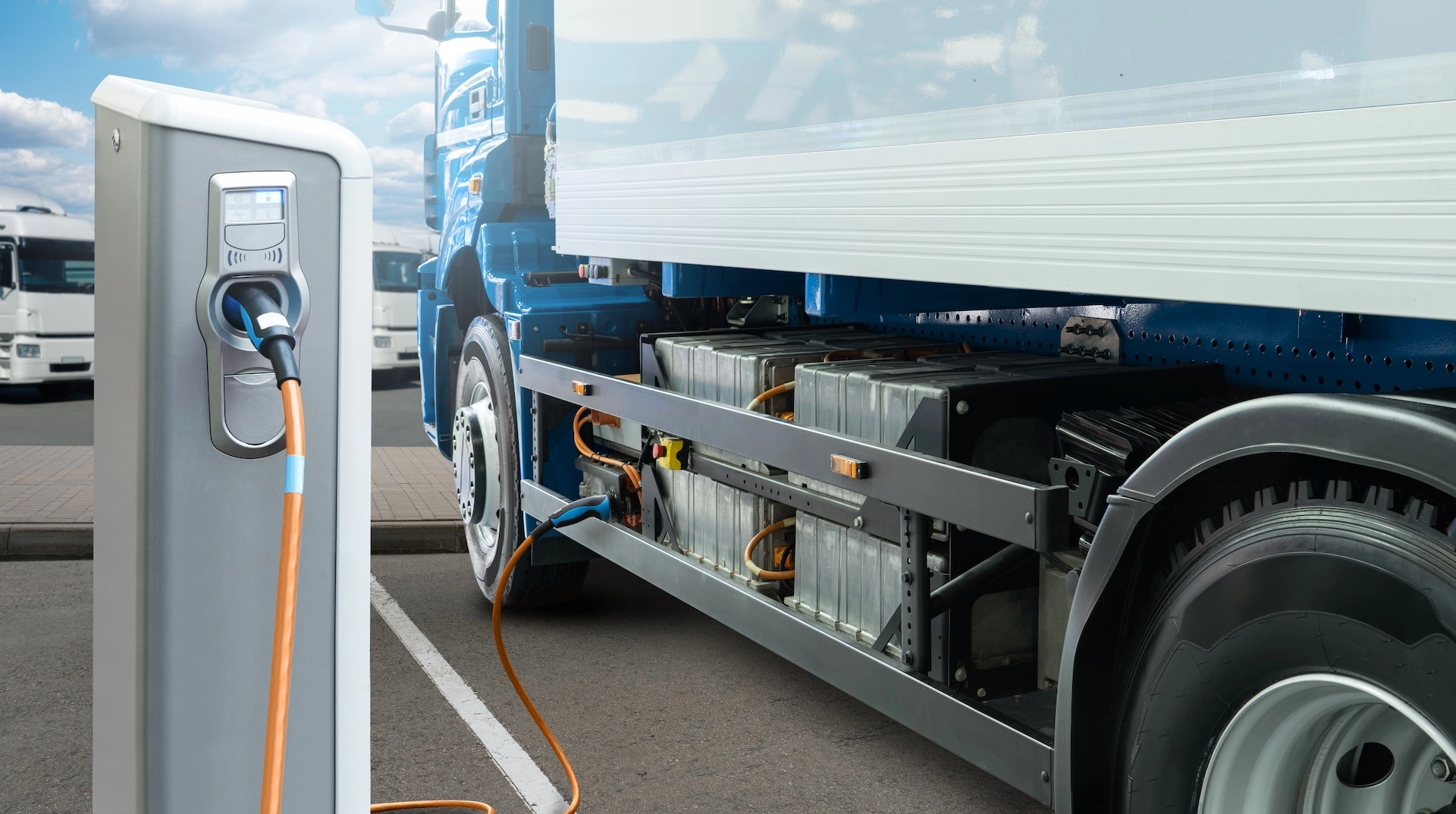 An electric truck charging at a depot, with its battery pack visible beneath the chassis.