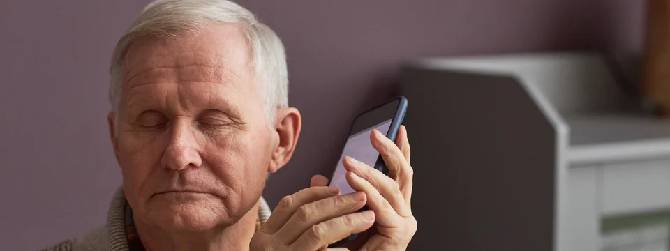 An older man with his eyes closed, holding a smartphone close to his face, listening rather than looking at the screen.