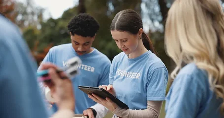 Charity volunteers in blue t-shirts working together outdoors, one using a tablet while others look on.
