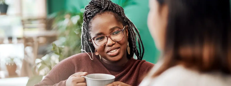 Two women sitting together and talking over mugs of tea or coffee, with one woman smiling warmly and actively engaging in the conversation in a cozy, indoor setting.