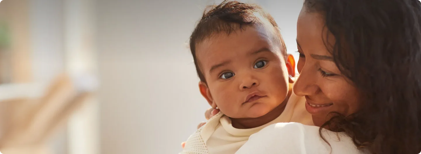 A mother smiles warmly while holding her baby, who looks directly at the camera with a calm expression.