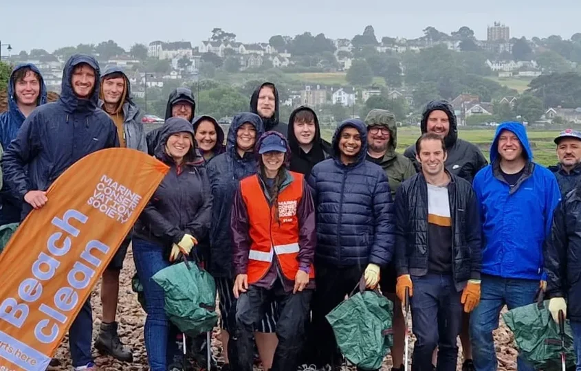 Group of Giant Digital team members smiling and posing together in rain gear during a beach clean event, holding litter pickers and an orange Marine Conservation Society banner.