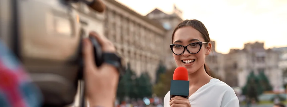 A female TV reporter holding a microphone and speaking to camera on a city street.