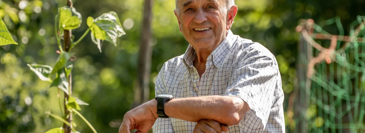 An elderly man smiling and leaning on a gardening tool in a lush, green garden on a sunny day.