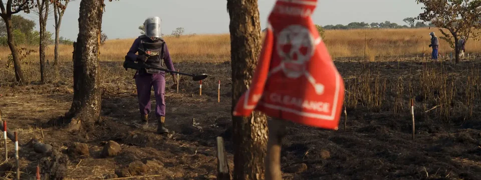 A deminer wearing protective gear and carrying detection equipment walks through a marked minefield, with a warning sign in the foreground and others working in the background.