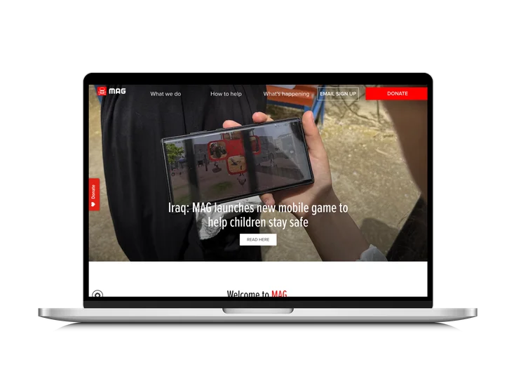 A laptop showing the Mines Advisory Group homepage, with "Iraq: MAG launches new mobile game to help children stay safe" headline over a photo of a hand holding a phone showing the game.