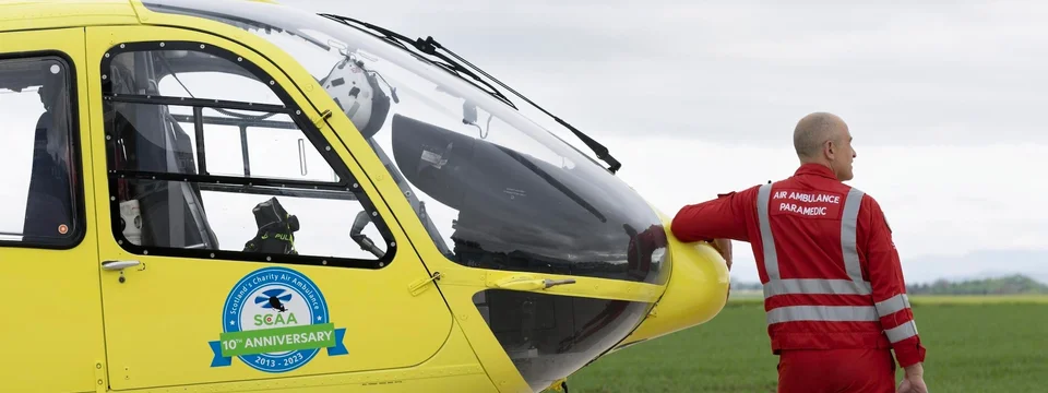 A paramedic in a red Air Ambulance uniform standing beside Scotland's Charity Air Ambulance helicopter, marked with a 10th Anniversary badge, on an airfield.