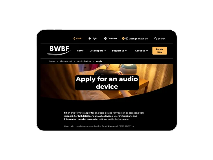 A tablet showing the British Wireless for the Blind Fund "Apply for an audio device" page in dark mode, with a form intro and a photo of an audio device.