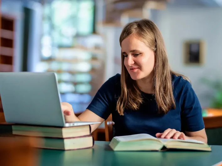 A visually impaired woman uses a laptop with large print books in a library setting, demonstrating accessible learning and digital inclusion.
