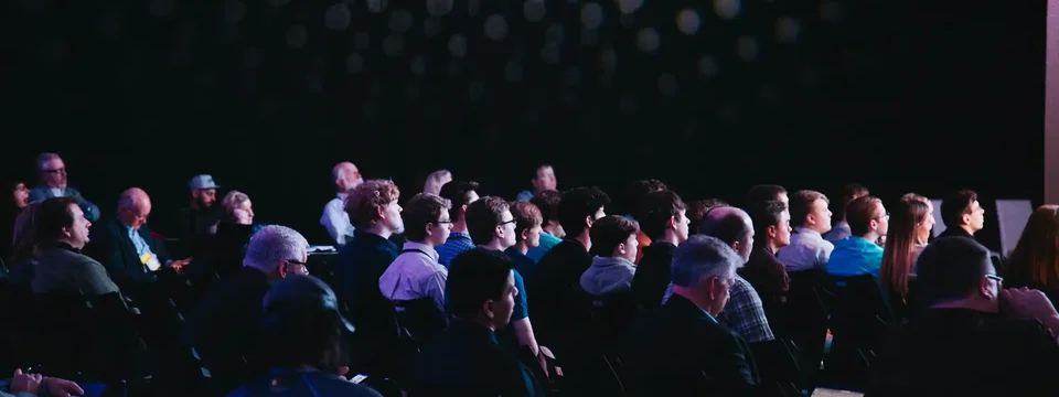 Audience seated in a dark auditorium, attentively watching a presentation or speaker under spotlights creating a patterned light effect on the wall.
