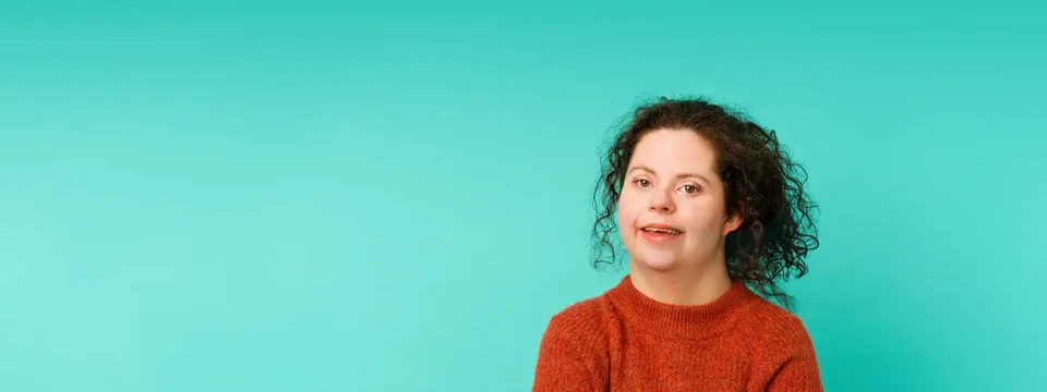 Young woman with curly hair wearing a red jumper standing confidently in front of a bright turquoise background, representing empowerment and inclusion.