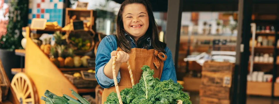 A smiling young woman wearing an apron holds a basket of fresh leafy greens and leeks, standing in front of a market stall filled with fruit and vegetables.
