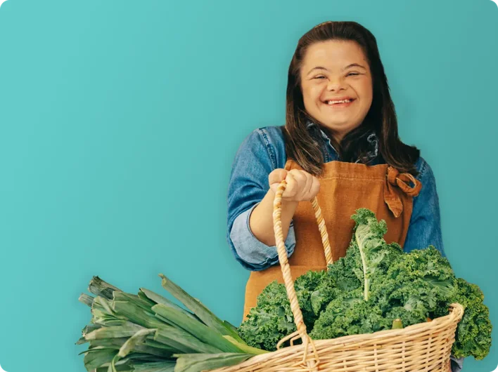 A smiling young woman wearing an apron holds a basket of fresh leafy greens and leeks, standing in front of a market stall filled with fruit and vegetables.