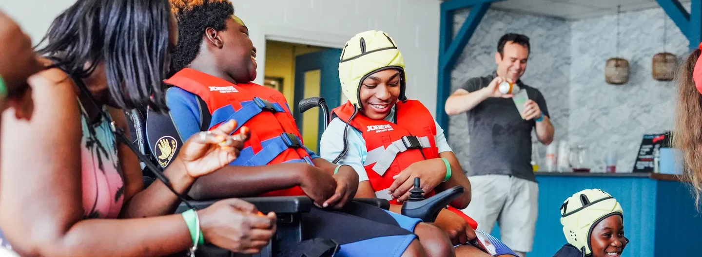 Two children in wheelchairs wearing red life jackets and smiling, one wearing a yellow protective helmet, interacting joyfully with an adult.