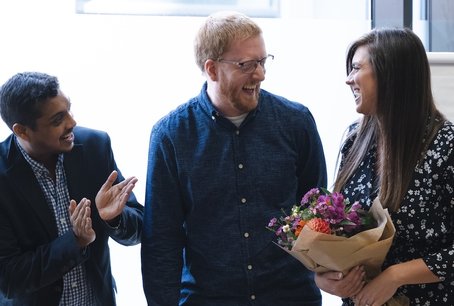 Three Giant Digital colleagues smiling and celebrating together in a bright office space. Kelly holds a bouquet of flowers, while Vim and Pete clap in appreciation.