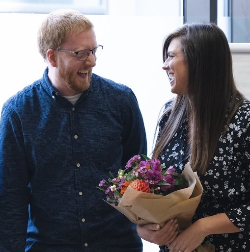 Three Giant Digital colleagues smiling and celebrating together in a bright office space. Kelly holds a bouquet of flowers, while Vim and Pete clap in appreciation.