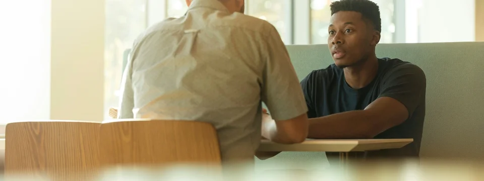 Two people talking together at a table near large windows, one facing the camera and speaking, the other seen from behind.