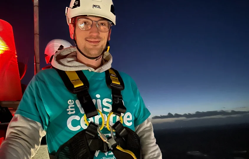 Tom, Senior Software Engineer, wearing a helmet and safety harness at height at dusk, wearing a Wish Centre charity T-shirt during an abseil fundraiser.