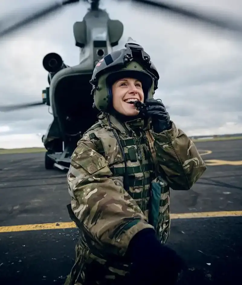 Smiling female soldier in camouflage uniform and helmet speaking into a radio, standing in front of a military helicopter on a tarmac.