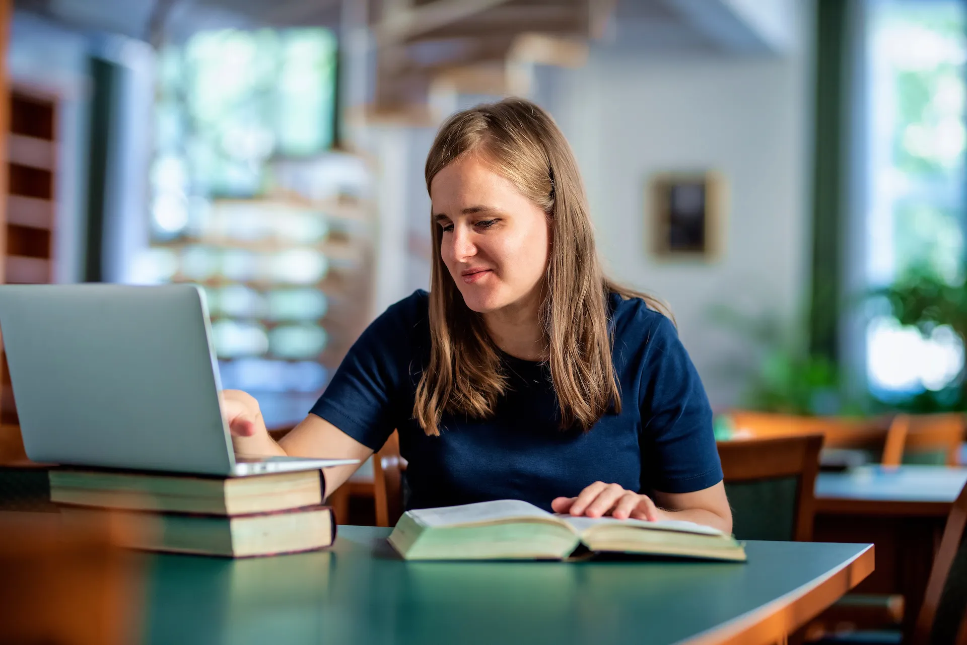 A visually impaired woman uses a laptop with large print books in a library setting, demonstrating accessible learning and digital inclusion.
