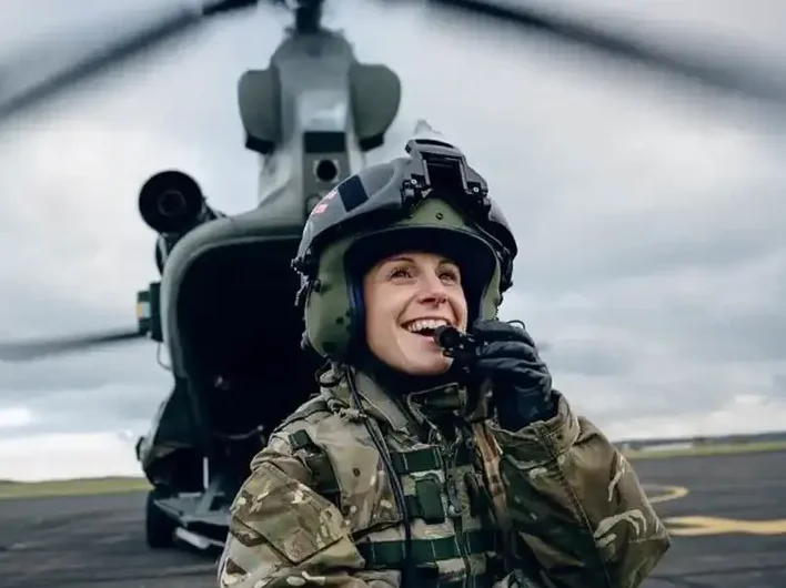 Smiling female soldier in camouflage uniform and helmet speaking into a radio, standing in front of a military helicopter on a tarmac.