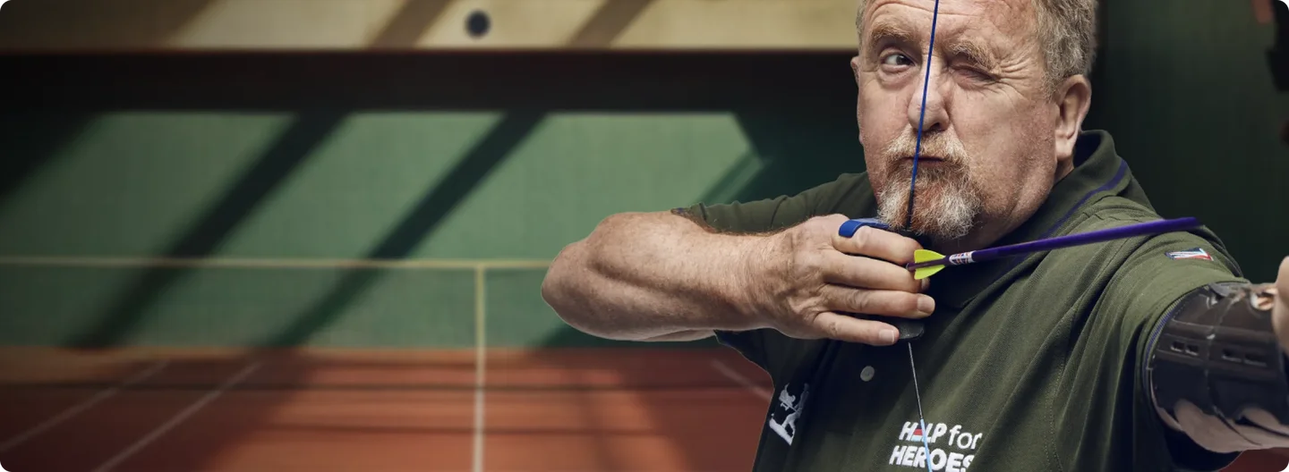A man in a green "Help for Heroes" shirt focuses intently as he aims a bow and arrow at an indoor archery range, demonstrating strength and concentration.