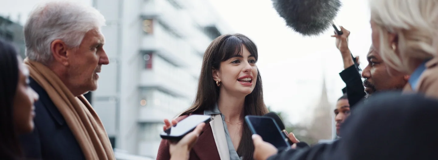A female interviewee surrounded by journalists holding microphones and phones for a street interview.