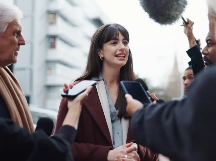 A female interviewee surrounded by journalists holding microphones and phones for a street interview.