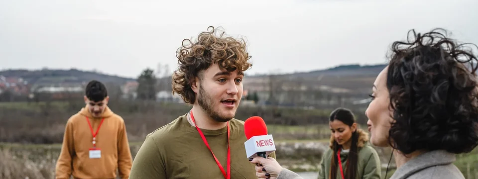 A reporter interviewing a volunteer with a "News" microphone, with other volunteers litter-picking in a field behind them.