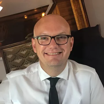 Peter is smiling on the Pullman train while celebrating his wedding anniversary, wearing glasses, a white shirt, and a black tie.