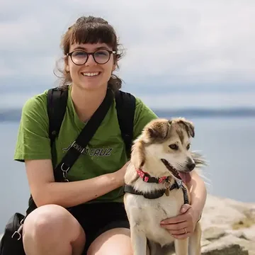 Gaby crouching by the seaside with her dog Maya, both looking happy and relaxed on a rocky outcrop with calm water behind.