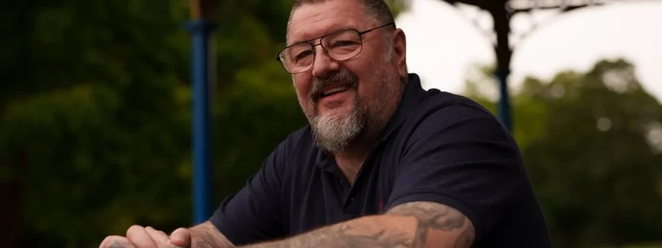 Smiling older man with glasses and tattoos on both arms, sitting outdoors under a shaded structure with greenery in the background.