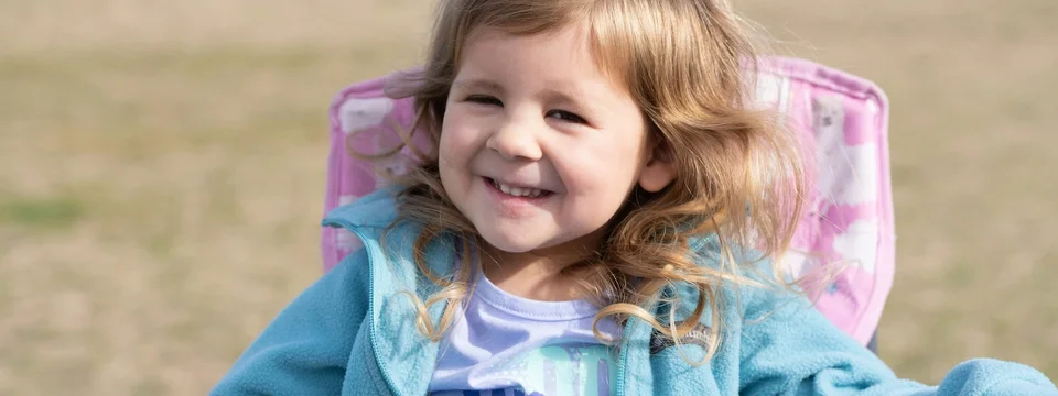 A young girl with curly hair smiling widely at the camera, wearing a blue fleece jacket, sitting in an outdoor mobility chair in a grassy field.