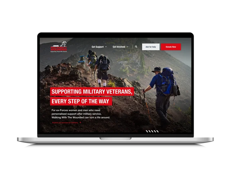A laptop showing the Walking With The Wounded homepage, with "Supporting military veterans, every step of the way" headline over a photo of hikers on a mountain trail.