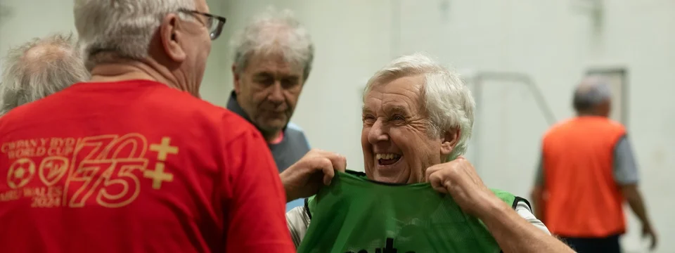 An older man laughing while pulling on a green 'Mitre' sports bib, with other men in red and orange kits standing nearby in an indoor sports hall.