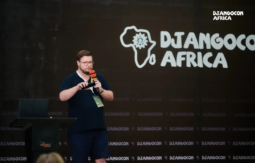 Jon, our Technical Director, speaking on stage at DjangoCon Africa, holding a microphone in front of a backdrop featuring the event’s logo and an outline of the African continent.