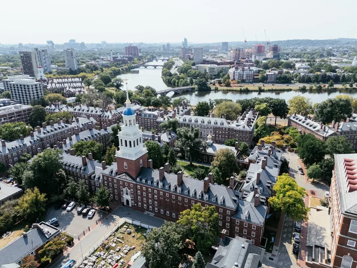 Aerial view of Harvard University's campus, with brick residential halls, a bell tower, and the Charles River winding through Cambridge beyond.