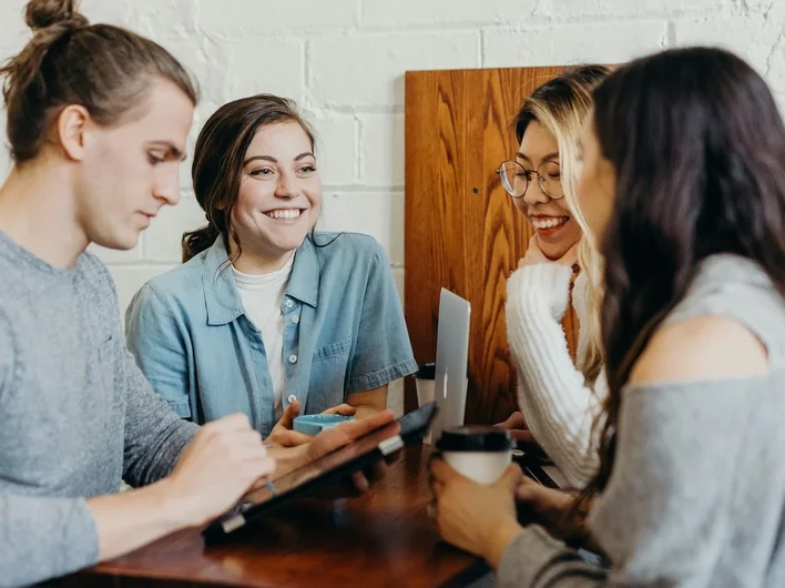 Four people sitting around a wooden table in a café, smiling and talking together, with laptops, a tablet, and coffee cups in front of them.