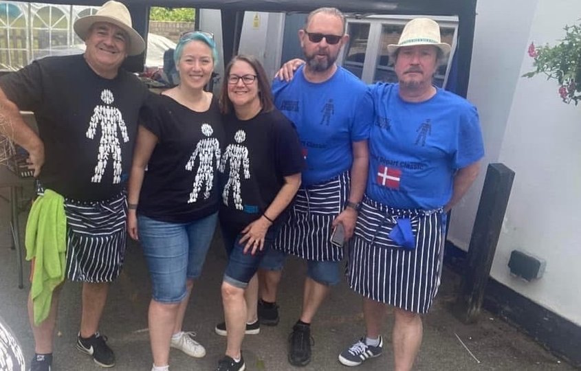 Mel, our Marketing Manager, with four friends wearing Prostate Cancer UK charity T-shirts and striped aprons at an outdoor fundraising event.