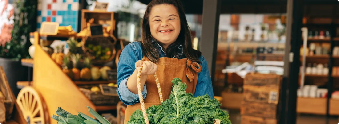 A smiling young woman wearing an apron holds a basket of fresh leafy greens and leeks, standing in front of a market stall filled with fruit and vegetables.