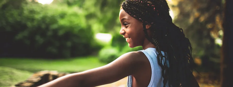 A young girl with braided hair smiles joyfully as she plays outdoors, arms outstretched in a moment of freedom and happiness. Sunlight filters through the trees behind her.