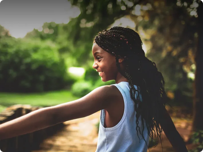A young girl with braided hair smiles joyfully as she plays outdoors, arms outstretched in a moment of freedom and happiness. Sunlight filters through the trees behind her.
