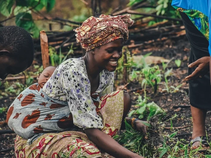 woman smiling while working the soil in a garden, carrying a sleeping baby wrapped on her back, with another person crouched nearby.