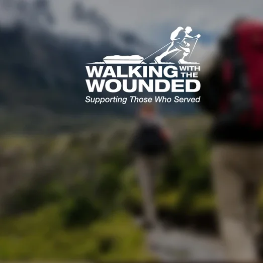 Walking with the Wounded logo over a blurred photo of two hikers with backpacks in a mountainous landscape.