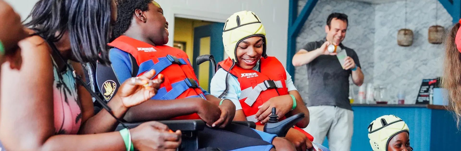 Two children in wheelchairs wearing red life jackets and smiling, one wearing a yellow protective helmet, interacting joyfully with an adult.