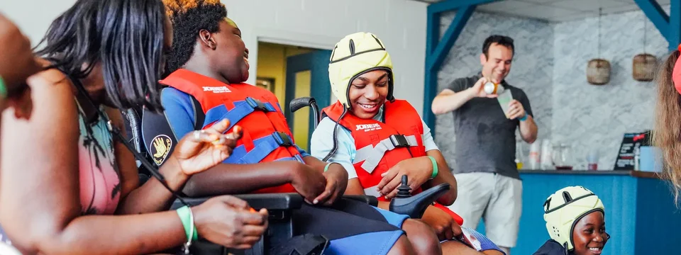 Two children in wheelchairs wearing red life jackets and smiling, one wearing a yellow protective helmet, interacting joyfully with an adult.