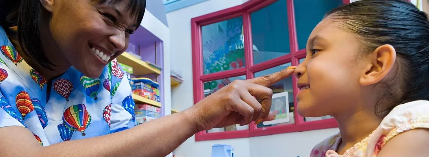 A smiling nurse gently touches a young girl’s nose while holding her arm, creating a warm, caring moment in a brightly coloured playroom.